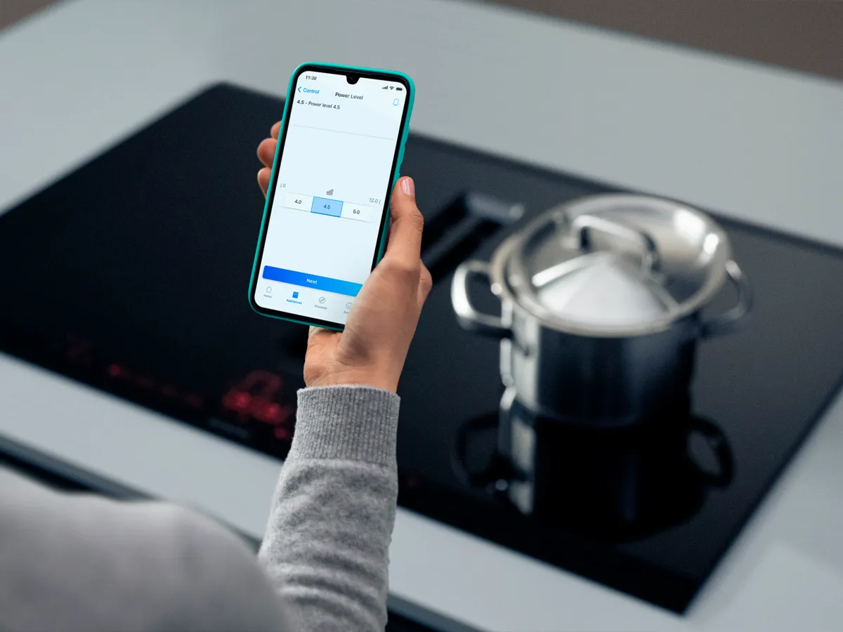 Pessoa que utiliza um smartphone para controlar um fogão de indução moderno com uma panela em cima.