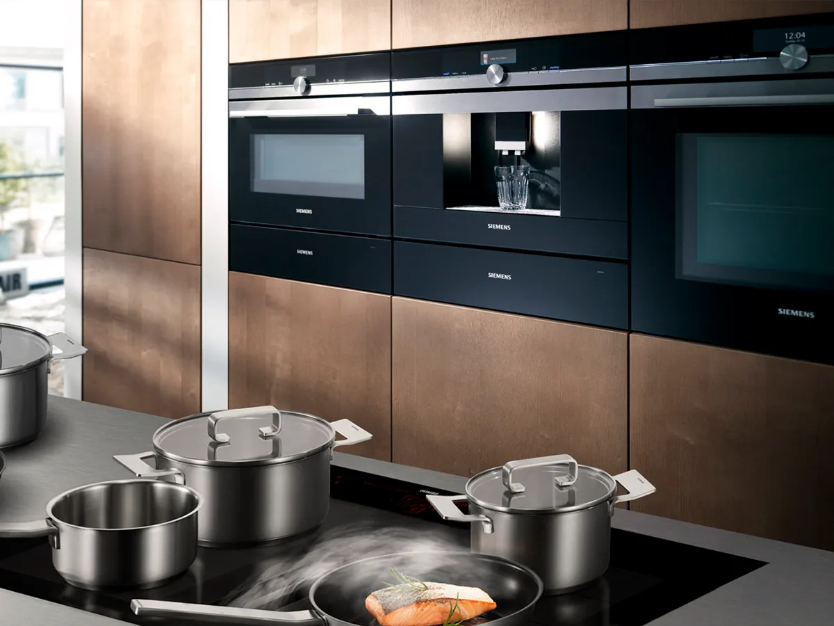 Placa de indução Siemens com vários tachos e uma panela de cozer a vapor, em contraste com elegantes electrodomésticos de cozinha encastrados.