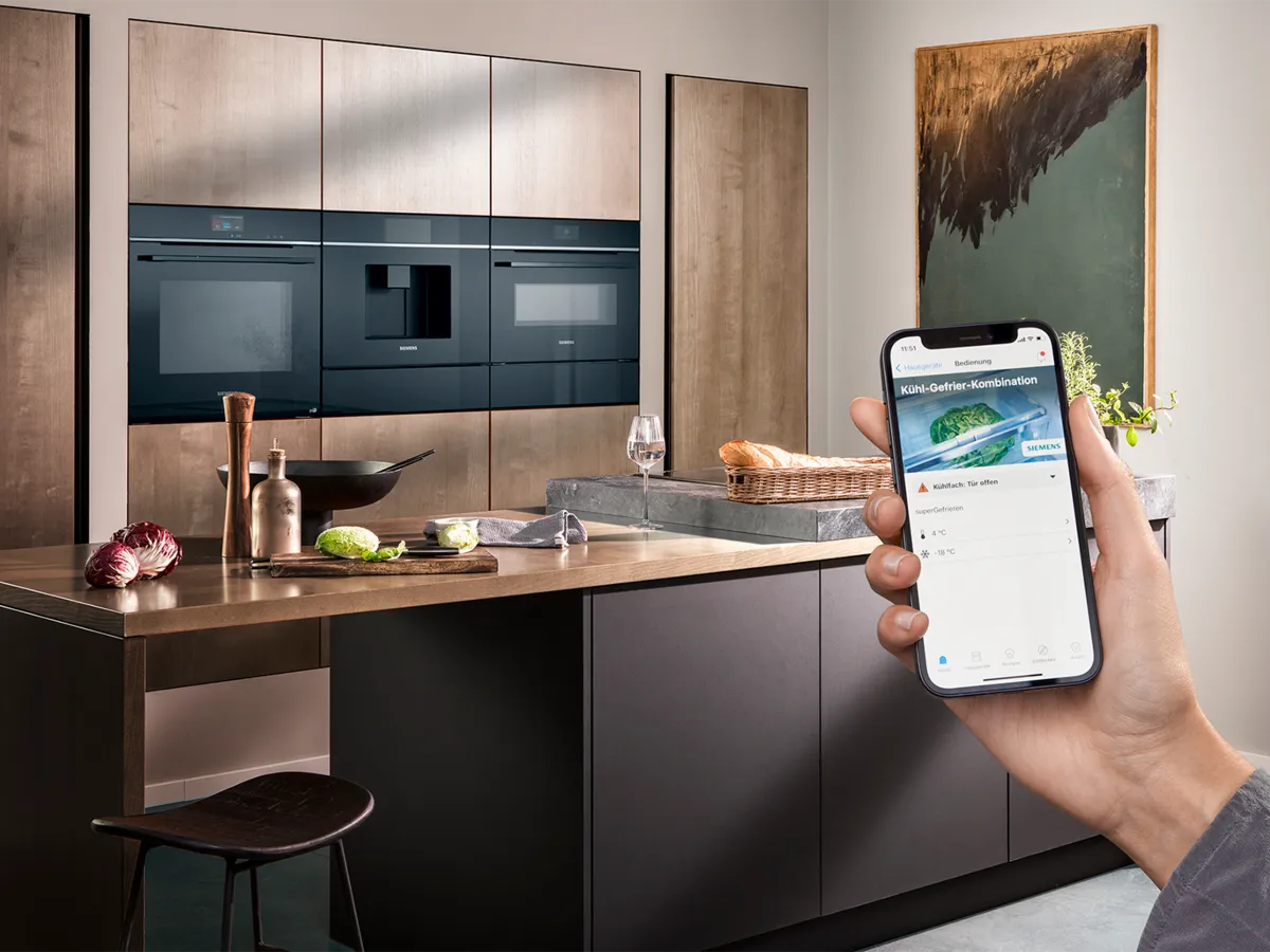 Mão segurando um smartphone com a aplicação Siemens Home Connect, controlando remotamente os aparelhos de cozinha incorporados.