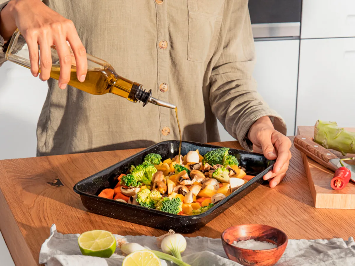 Uma pessoa regando azeite sobre uma bandeja de legumes frescos, preparando uma refeição caseira em um ambiente de cozinha moderna.