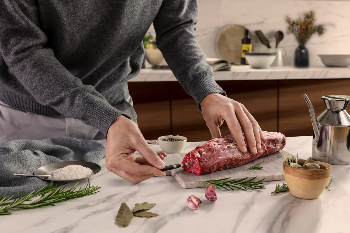 A person in a grey sweater prepares a raw cut of meat on a marble countertop by inserting NEFF meatprobe.