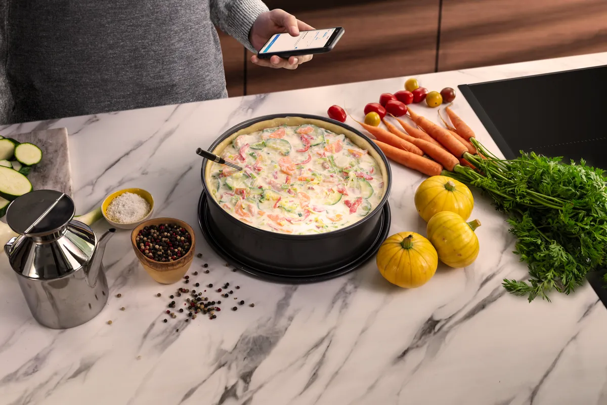 A quiche in a round black pan, surrounded by colorful fresh vegetables, spices, and a stainless steel oil dispenser.