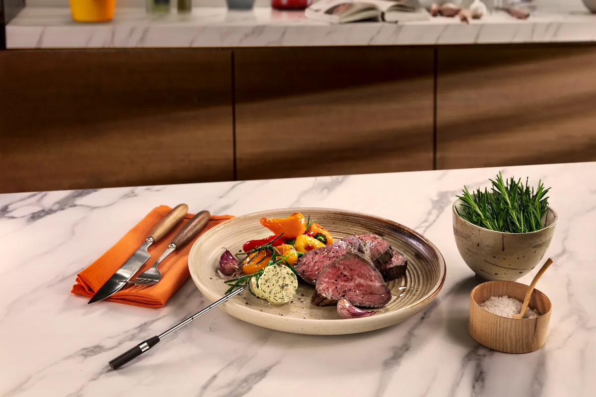Plated gourmet steak with garlic butter, roasted vegetables and herbs, accompanied by cutlery.
