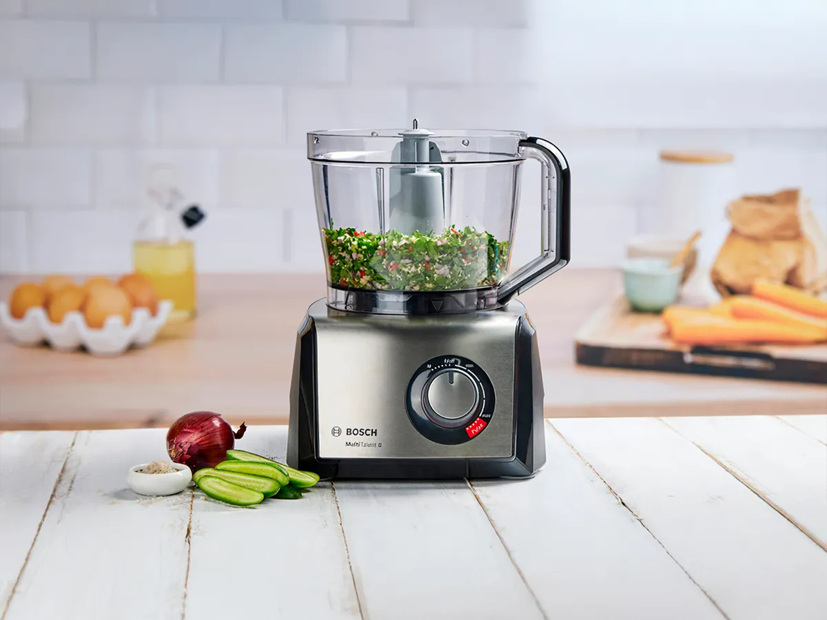 Processador de alimentos Bosch sobre balcão de madeira, rodeado de legumes frescos, pronto para a preparação de refeições.