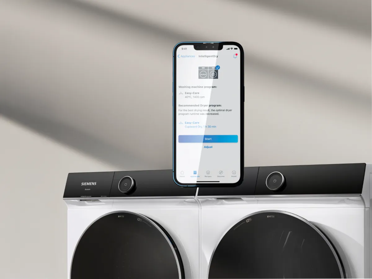 Um smartphone a mostrar uma aplicação de lavandaria colocado em cima de uma máquina de lavar Siemens, com uma secadora correspondente ao lado.
