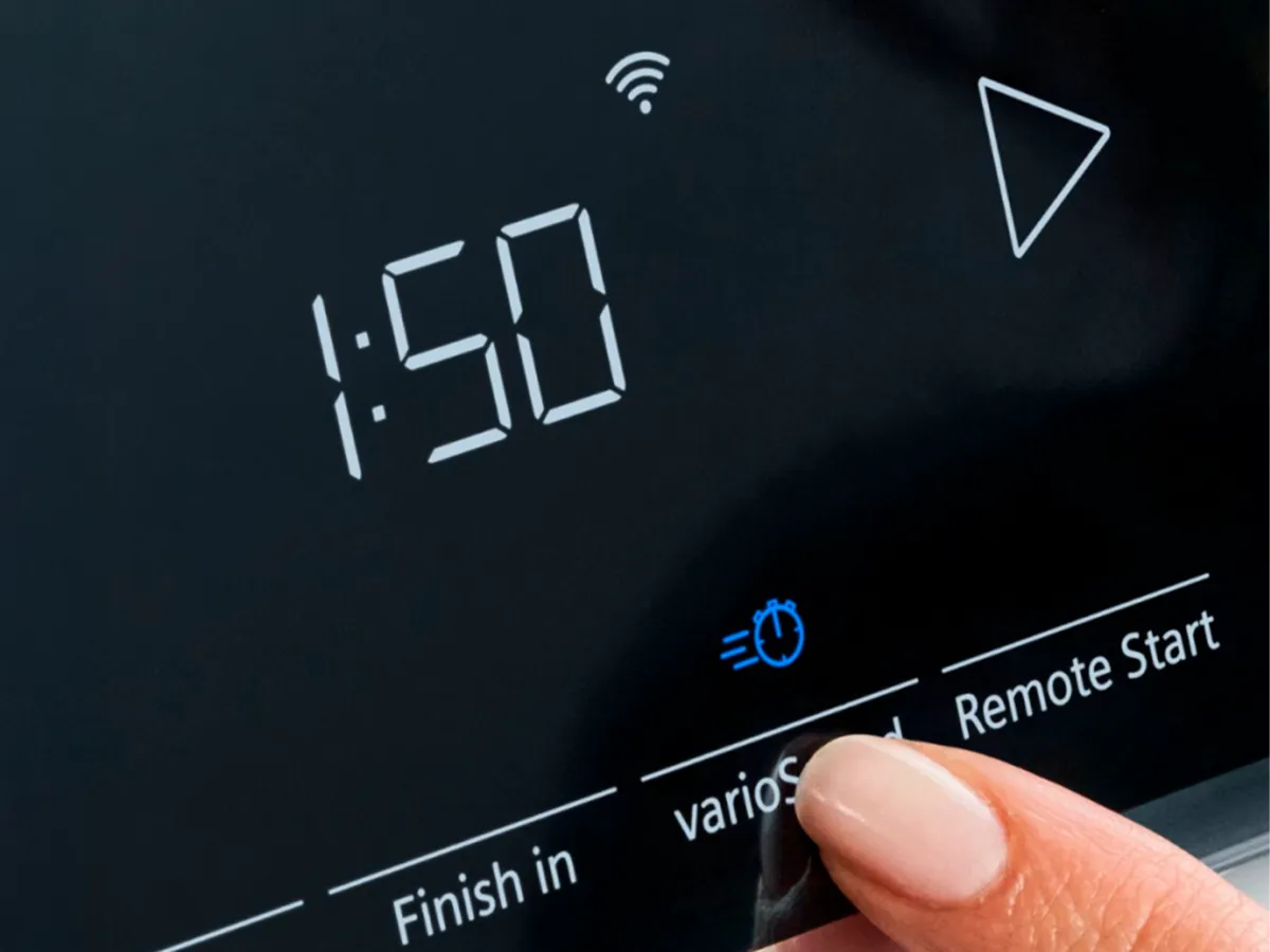Ecrã digital a mostrar 1:50 com um dedo a pressionar um botão iluminado e ícones de Wi-Fi e início remoto.