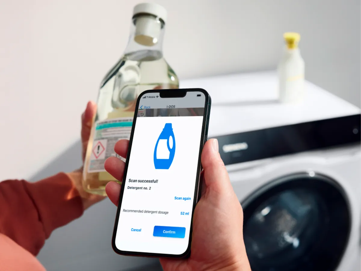 Mão a segurar um smartphone com a aplicação a escanear uma garrafa de detergente perto da máquina de lavar.