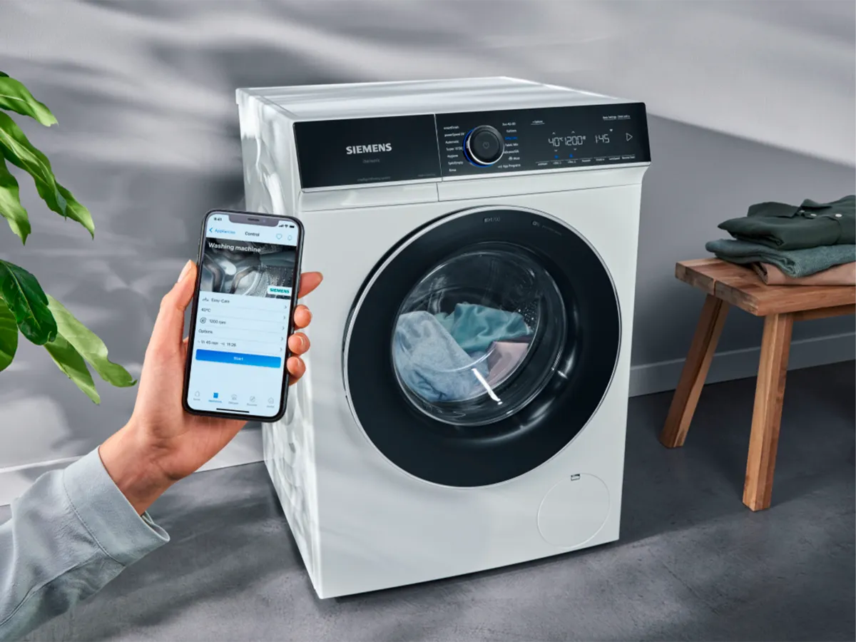 Mão a segurar um smartphone com uma aplicação aberta em frente a uma máquina de lavar moderna.