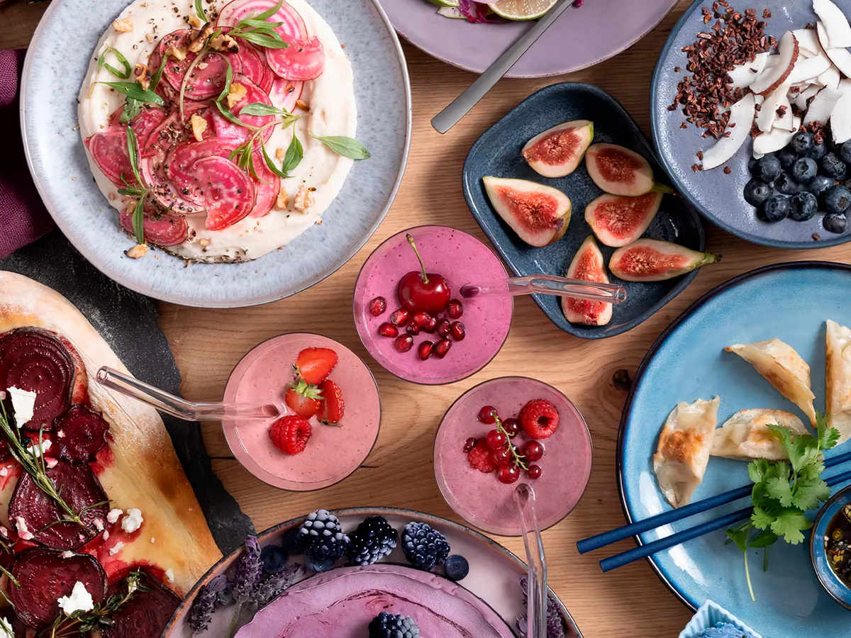 A colorful spread of smoothies, fruits, dumplings, beet dishes, and garnishes on a wooden table