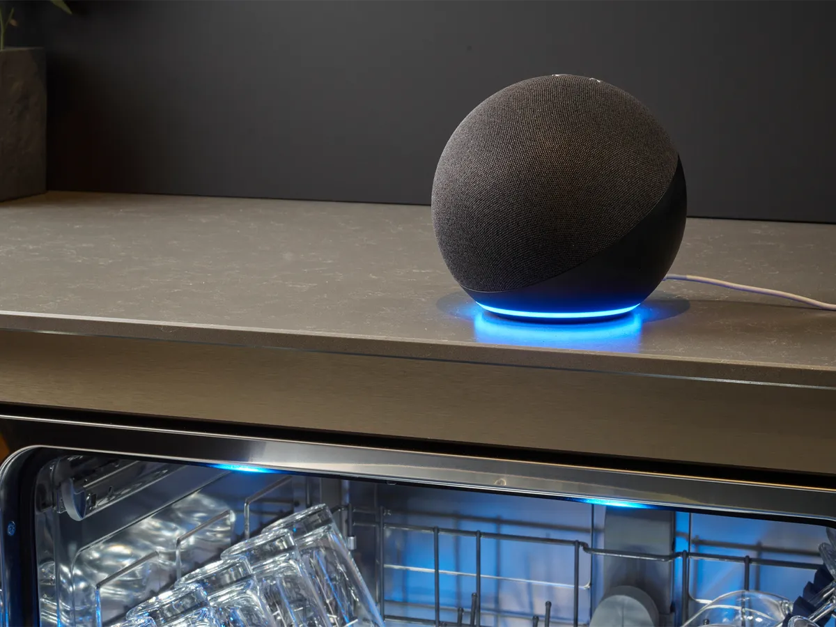 Echo Dot sobre uma bancada de cozinha com uma base azul iluminada, acima de uma máquina de lavar louça aberta.