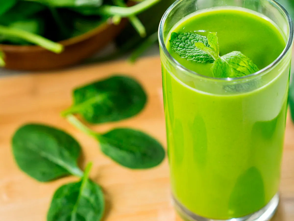 Um smoothie verde de abacate em pé sobre uma mesa, com folhas de espinafre ao lado.