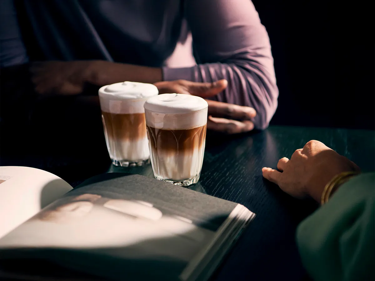 To personer sidder ved et bord med lagdelte kaffe kaffedrikke, mens en åben bog ligger i forgrunden.