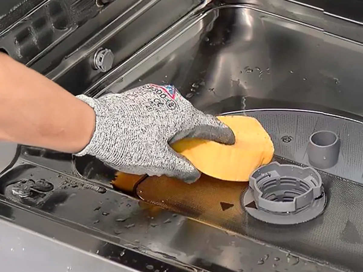 Gloved hand cleaning the filter inside a stainless steel dishwasher with a sponge.