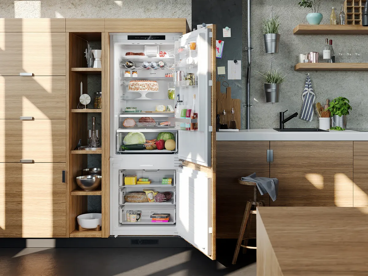 An open built-in fridge freezer inside a light-wood kitchen illuminated by daylight.