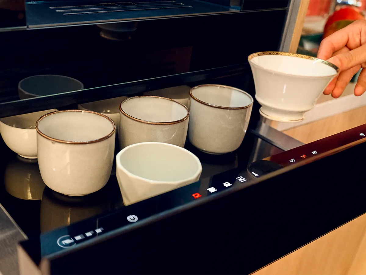 A closer view at an open Warming Drawer beneath an oven, filled with various cups, and a hand is removing a cup