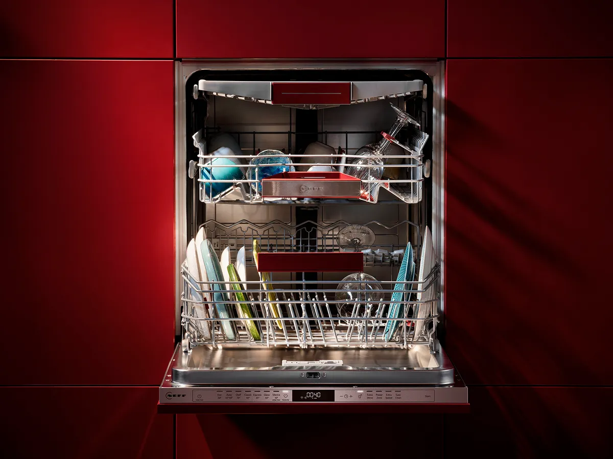 A modern dishwasher filled with clean plates, glasses, and colorful bowls, set against a deep red cabinet backdrop.