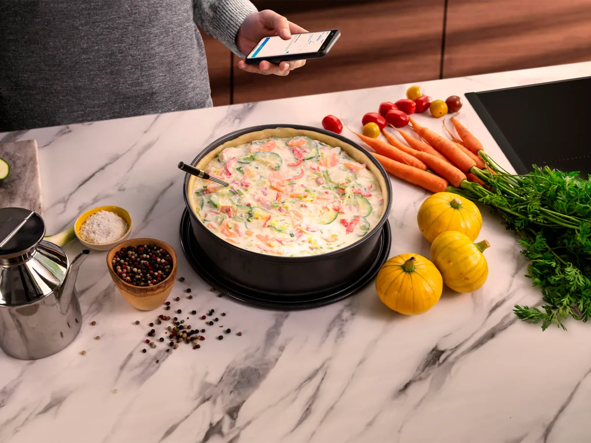 Lahtine pirukas ümmargusel mustal pannil, mida ümbritsevad värvilised värsked köögiviljad, vürtsid ja roostevabast terasest õlikann.