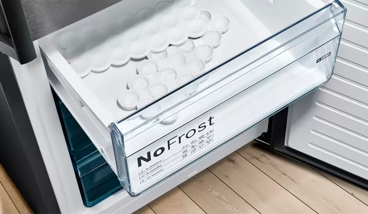 Un tiroir de congélateur transparent étiqueté « NoFrost » contient plusieurs bacs à glaçons remplis de glace, avec un intérieur organisé et des appareils modernes en arrière-plan.