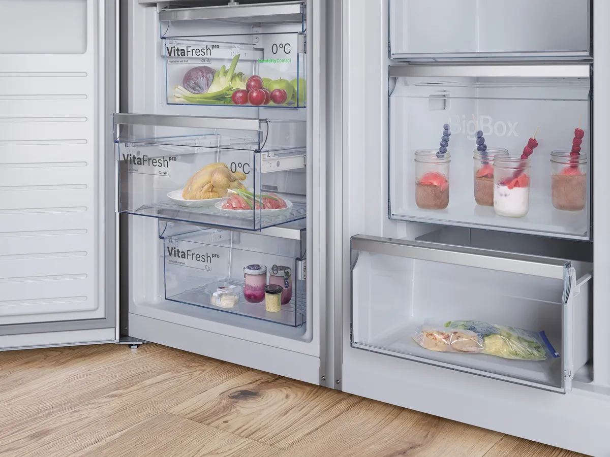Interior de um frigorífico moderno a mostrar alimentos frescos em gavetas transparentes sobre um piso de madeira.