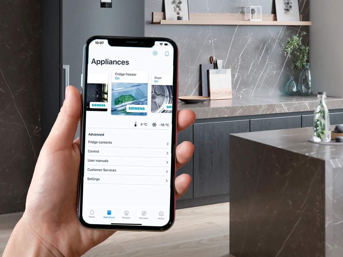Uma mão a segurar um smartphone que exibe uma aplicação de gestão de aparelhos numa cozinha moderna com superfícies de mármore cinza e um frigorífico elegante.