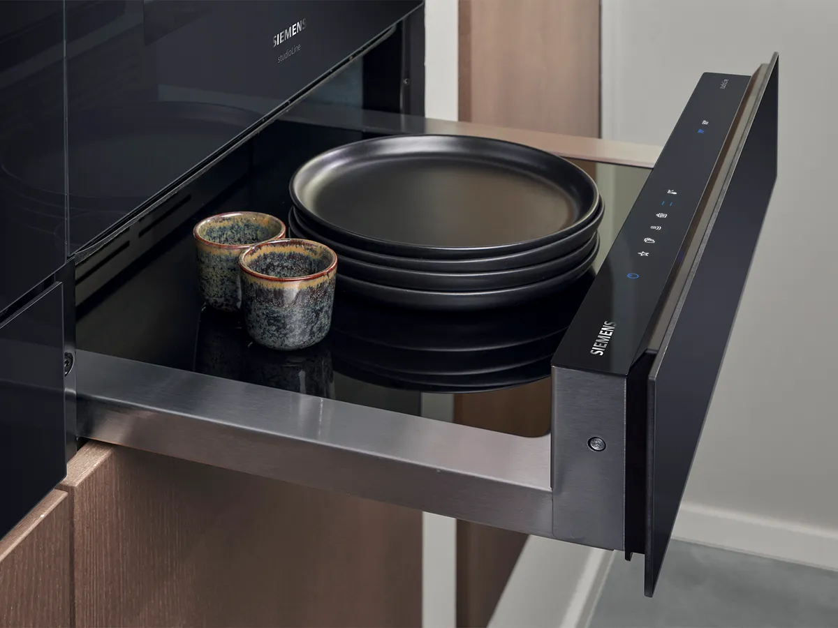 Siemens built-in warming drawer extended, holding stacked plates beneath a sleek oven for seamless kitchen integration.