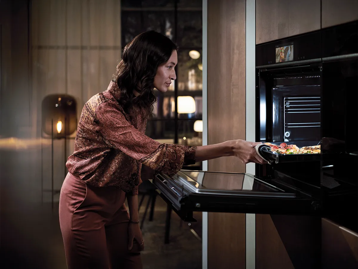 Uma mulher com um traje elegante entra num forno moderno, retirando um tabuleiro de legumes coloridos numa cozinha bem iluminada.
