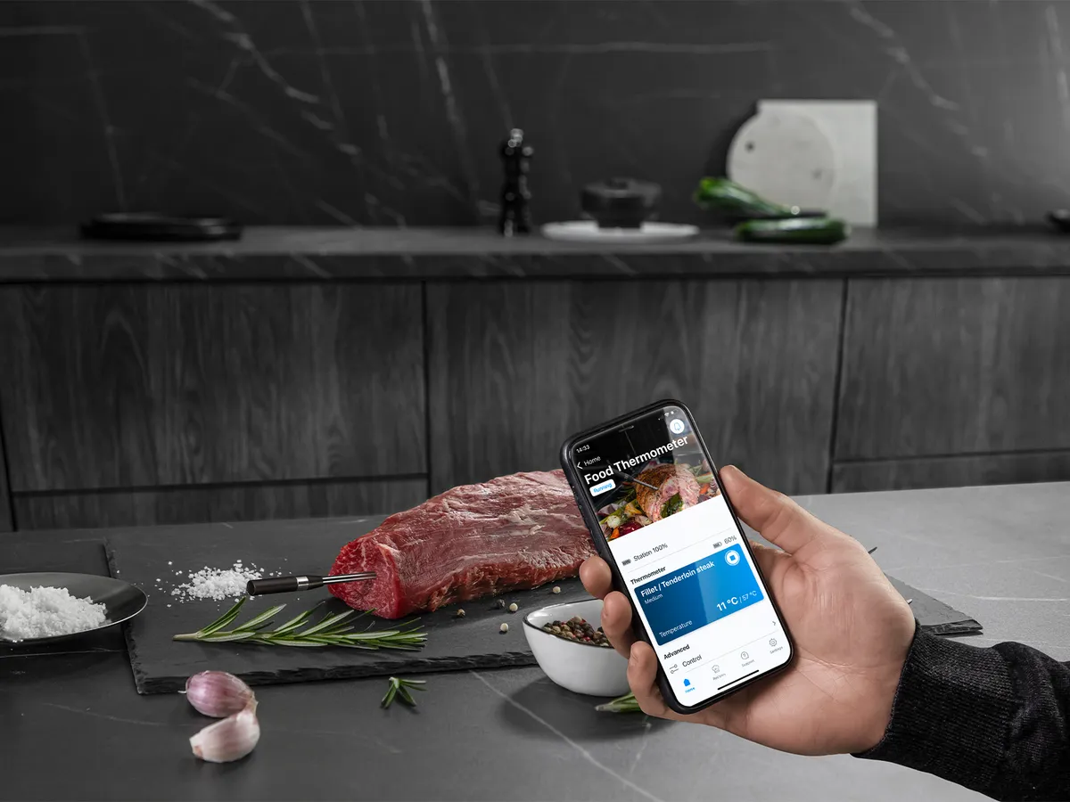 A person holds an iPhone while a steak rests on the counter, showcasing a wireless roasting sensor for cooking.  