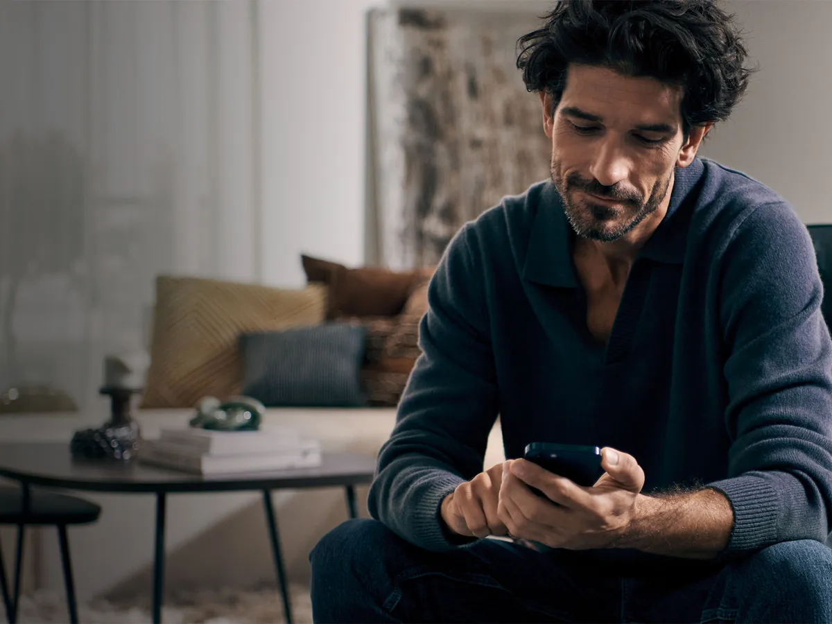 En man sitter på golvet i ett mysigt vardagsrum med en smartphone i handen, omgiven av mjuka textilier och dekorativa detaljer.