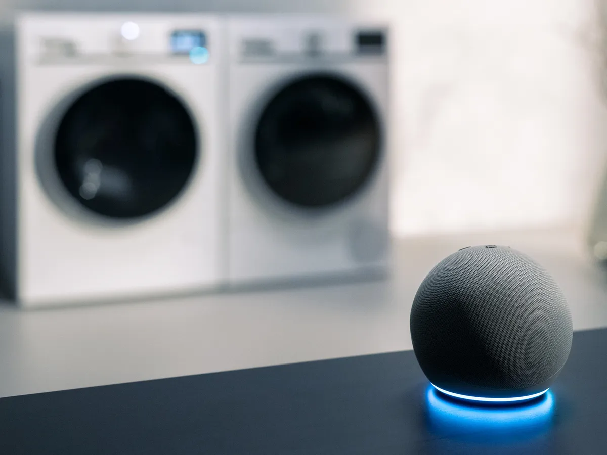A smart speaker with a blue light sits on a table, with washer-dryer units in the background.