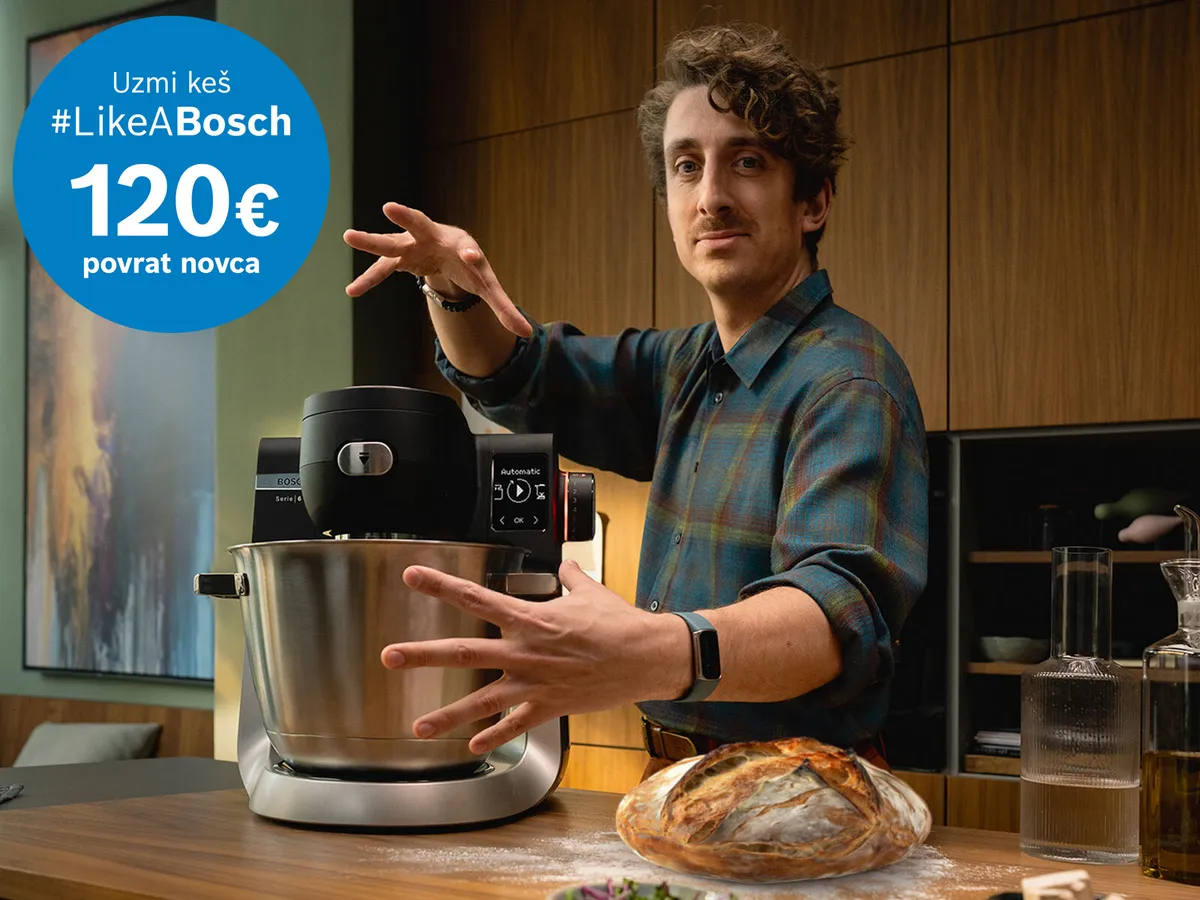 Osoba demonstrira Bosch kuhinjski mikser na drvenoj radnoj ploči, okružena staklenim posuđem i kruhom