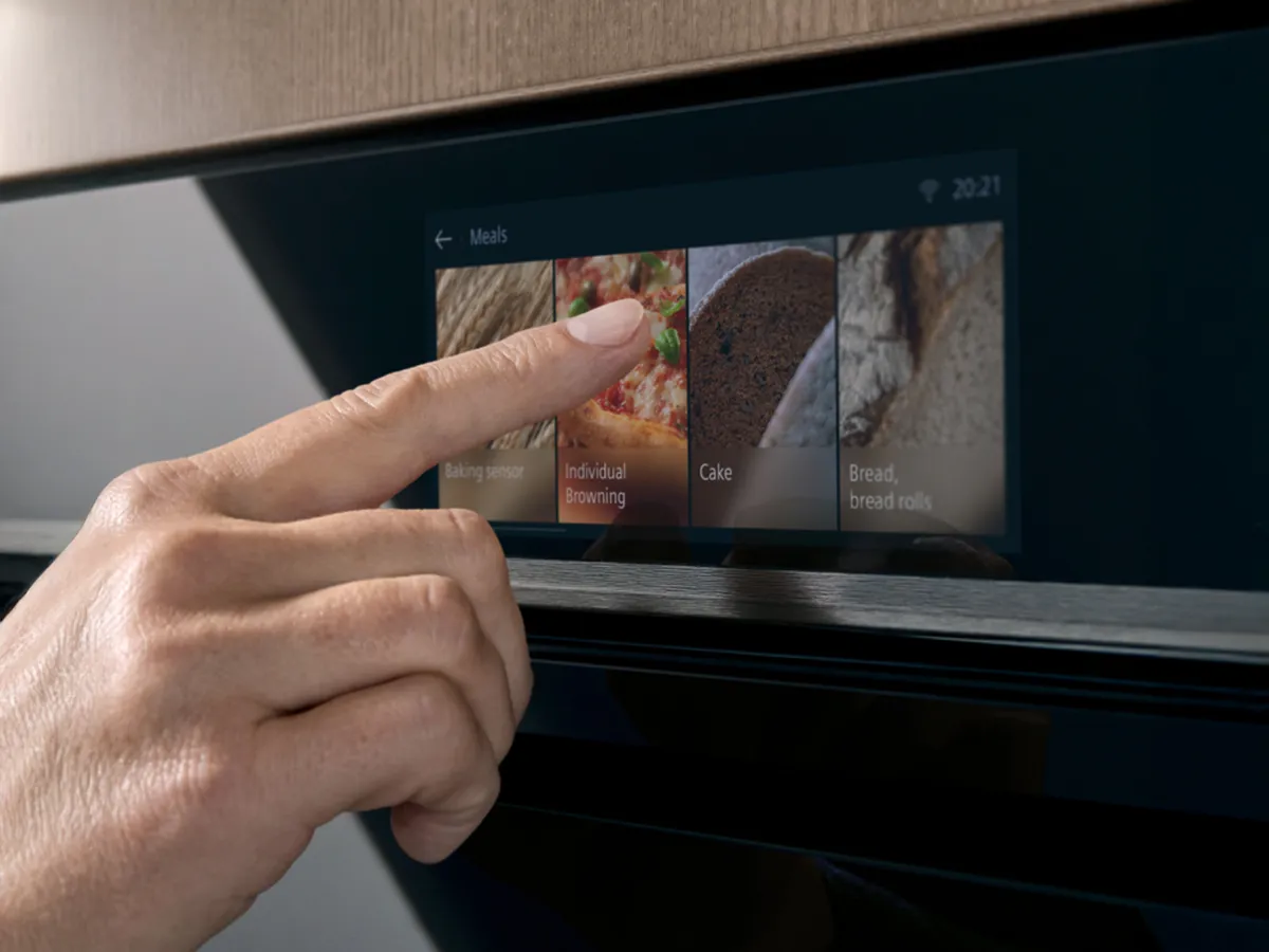 Uma mão interage com um painel de controlo do forno com ecrã tátil, selecionando opções de cozedura como “Bolo” e “Pãezinhos”.