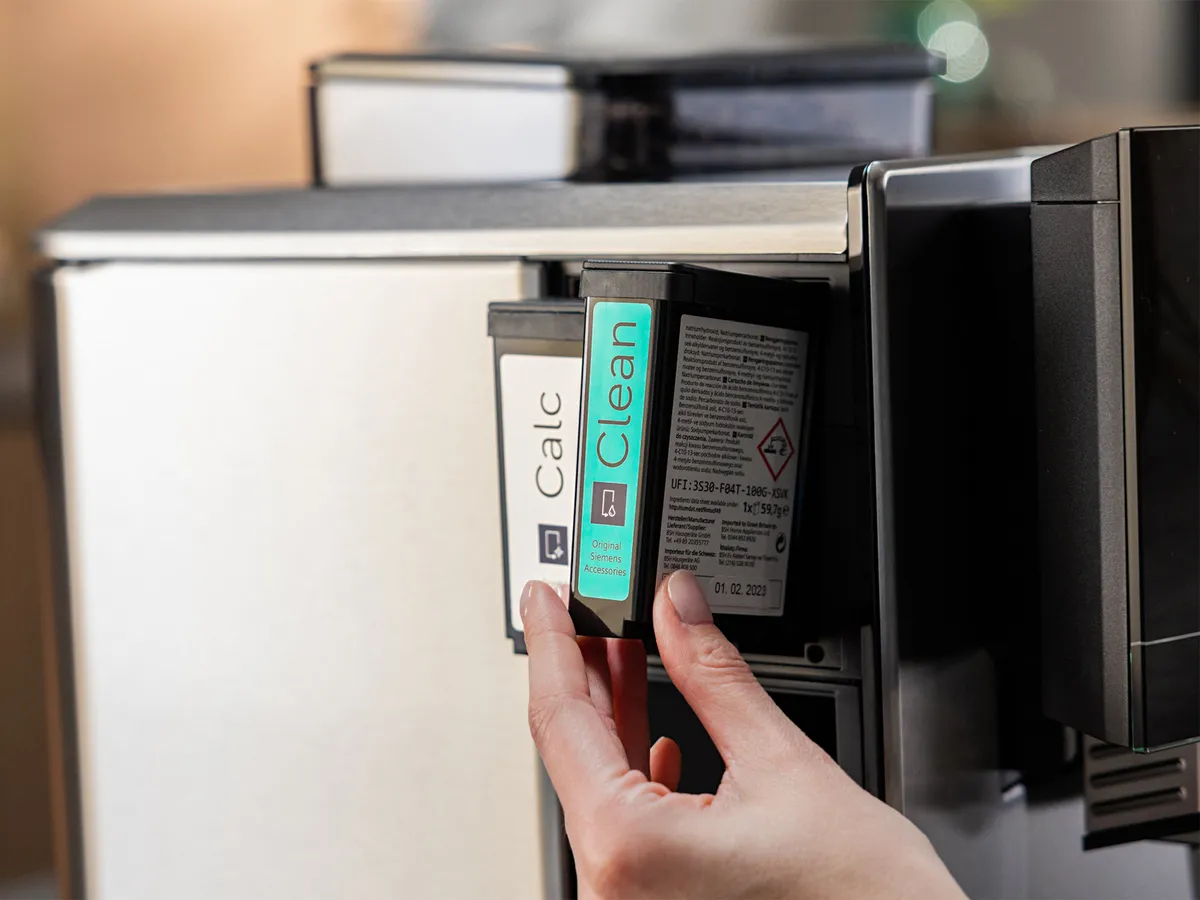 En persons hænder, der udskifter rengørings- og afkalkningspatroner på en Siemens EQ900 Plus fuldautomatisk kaffemaskine.