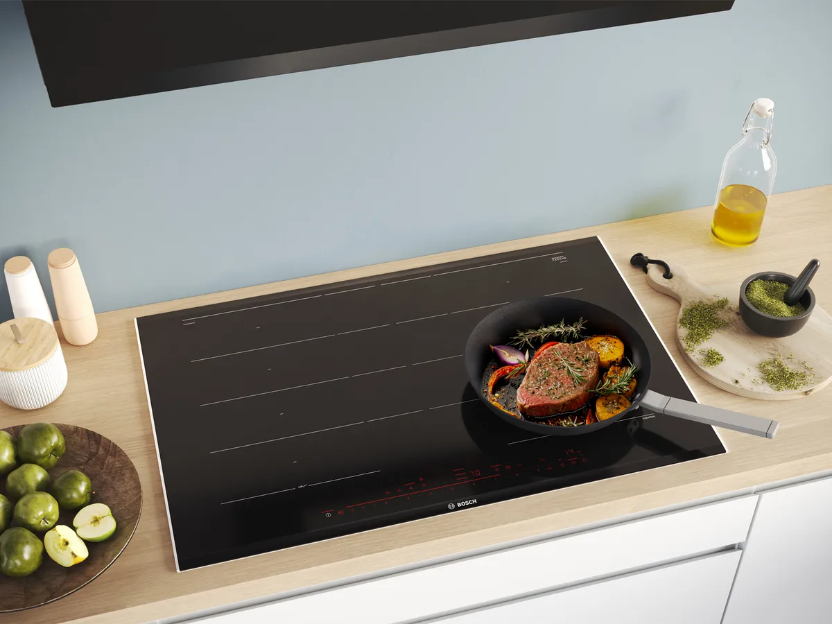 Placa de indução moderna com uma panela cozinhando legumes e carne, rodeada de utensílios de cozinha.