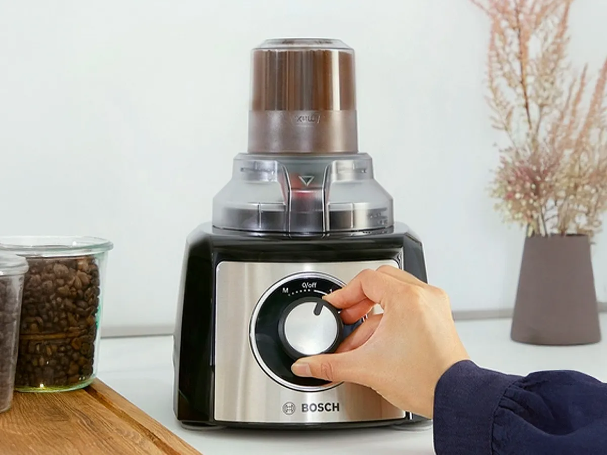 Uma pessoa ajusta o botão de um processador de alimentos Bosch num balcão, com grãos de café num frasco próximo.