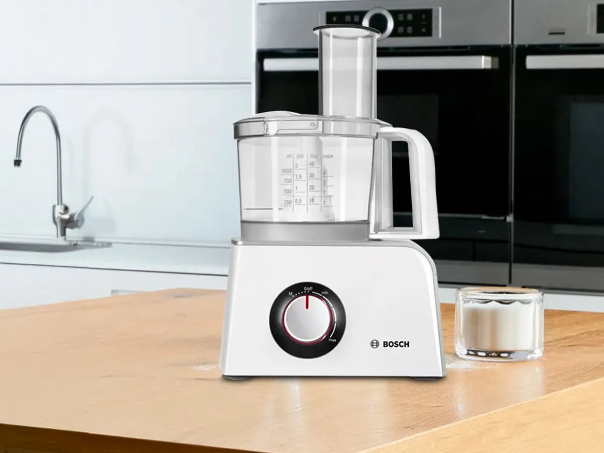 Um processador de alimentos Bosch branco num balcão de madeira, com um copo de açúcar ao lado.