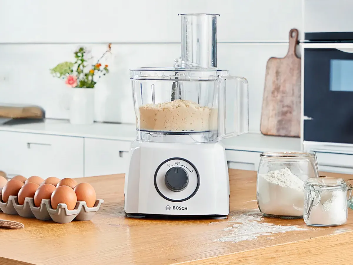 Um processador de alimentos Bosch num balcão de cozinha de madeira, rodeado de ovos e farinha.