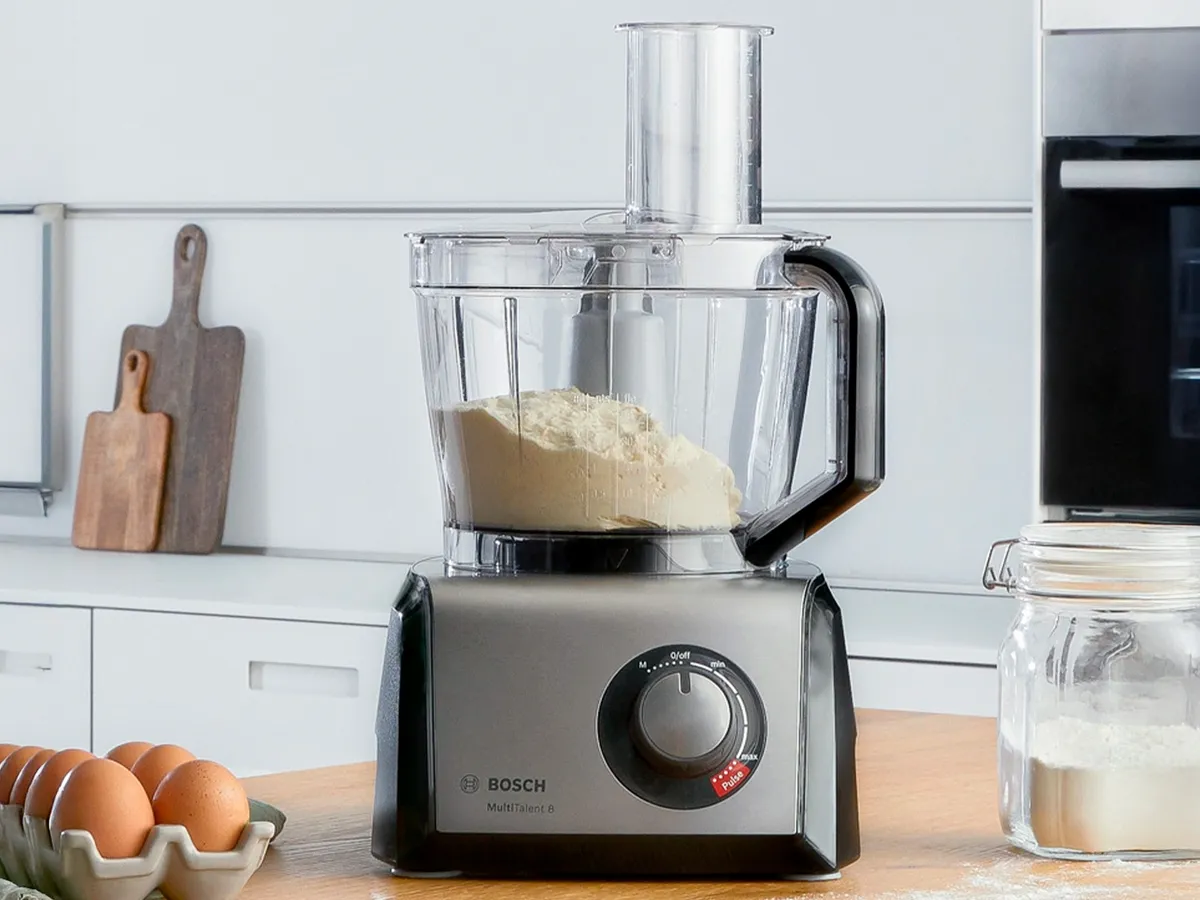 Um processador de alimentos Bosch numa mesa de cozinha de madeira com farinha dentro, ao lado de ovos numa tigela e um frasco de farinha.
