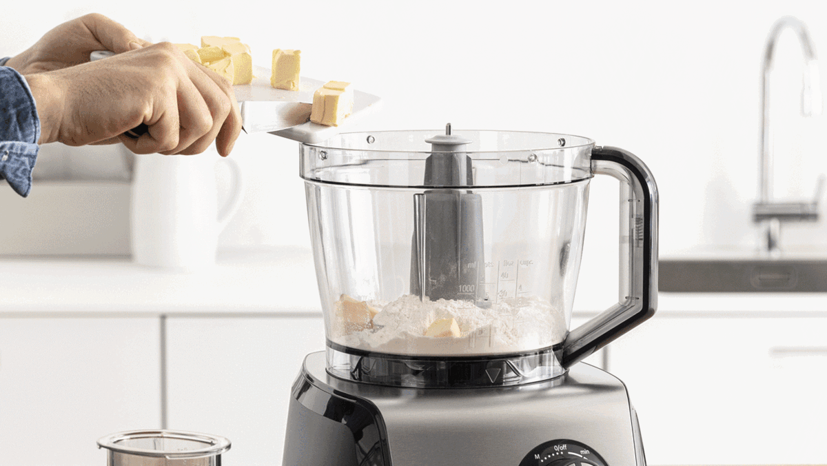 Uma pessoa a adicionar cubos de manteiga ao processador de alimentos para fazer massa.