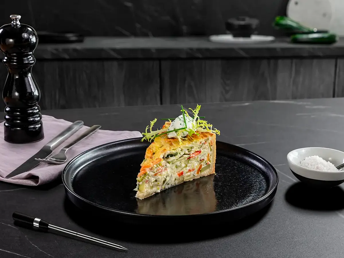 En skiva grönsaksquiche på en svart tallrik som ligger på en mörkgrå bänkskiva bredvid en pepparkvarn.