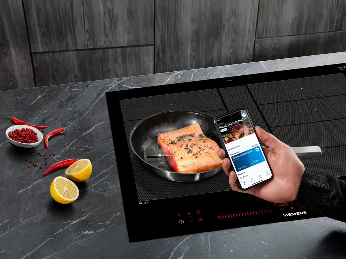 En hand som håller en smartphone framför lax i en panna med den trådlösa roastingSensor insatt.