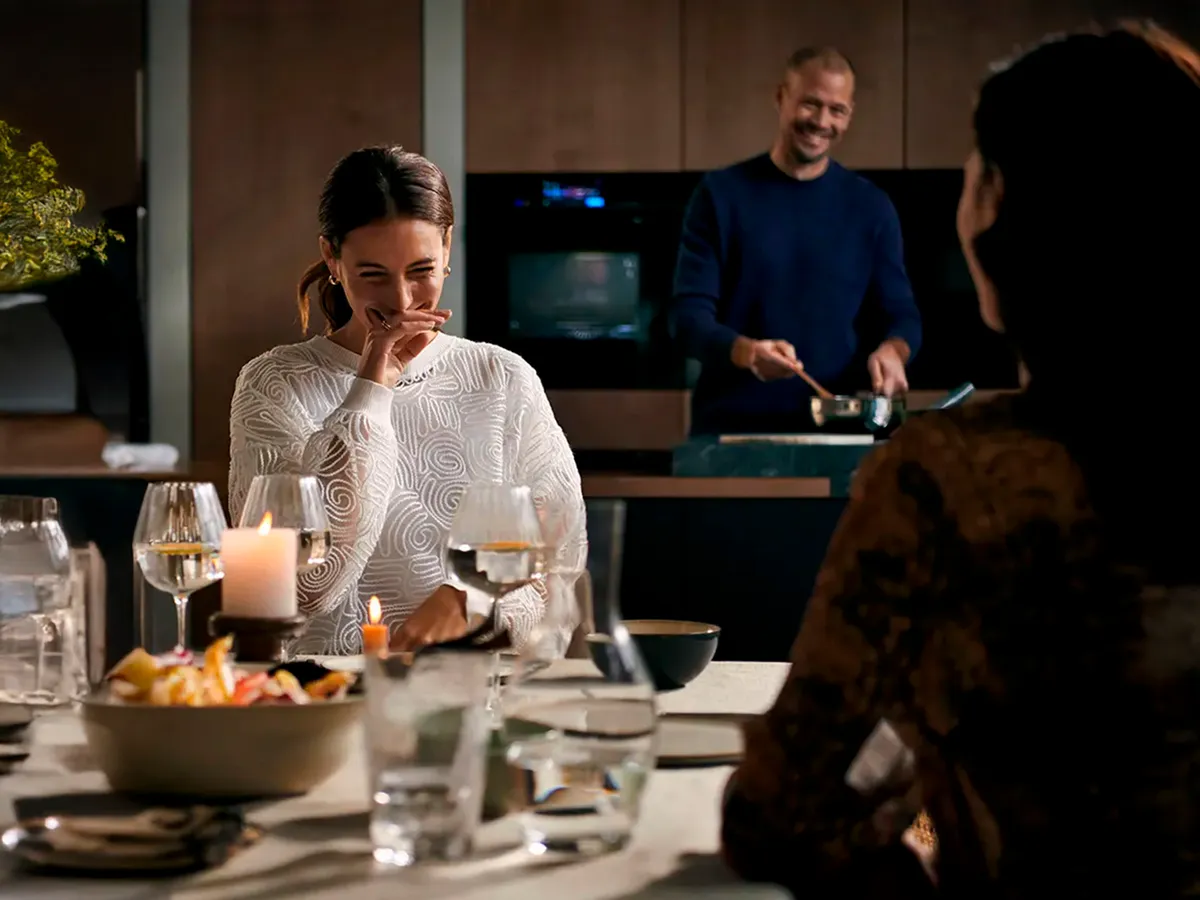 Två kvinnor sitter vid ett middagsbord medan en man serverar mat i köket i bakgrunden.