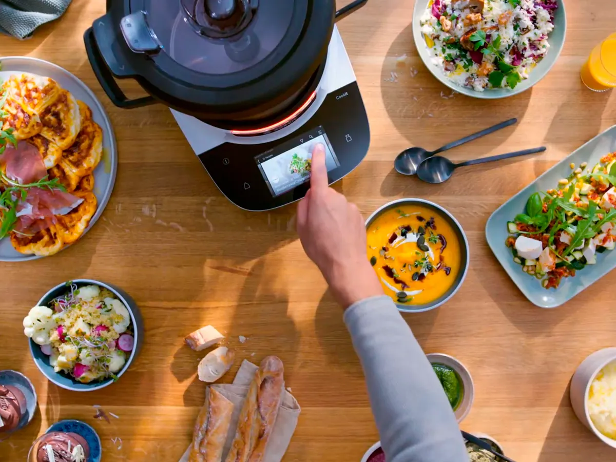 A hand operates a cooking device amidst a spread of dishes on a wooden table.
