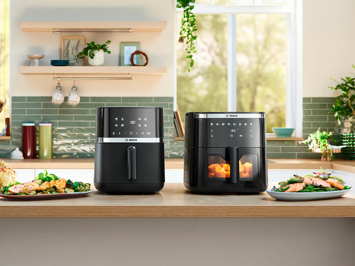 Dois modelos de fritadeiras a ar Bosch posicionados lado a lado em uma bancada de cozinha iluminada. Vários pratos e tigelas de comida, incluindo pão de ervas, frango, assados e batatas fritas, estão colocados ao lado dos eletrodomésticos, exibindo pratos feitos usando as fritadeiras a ar.