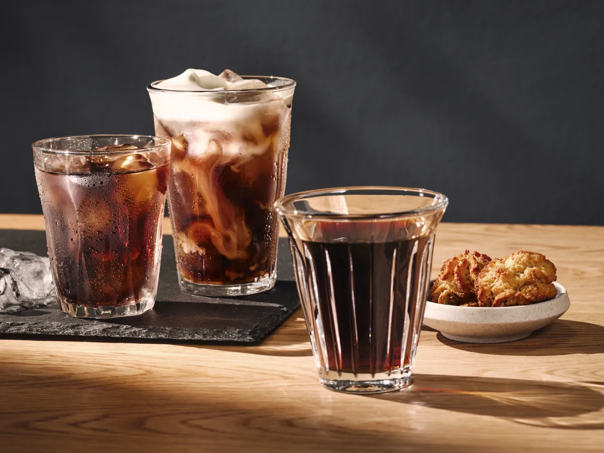 Tre glas iskold kaffe og en skål småkager på et træbord med isterninger i nærheden.