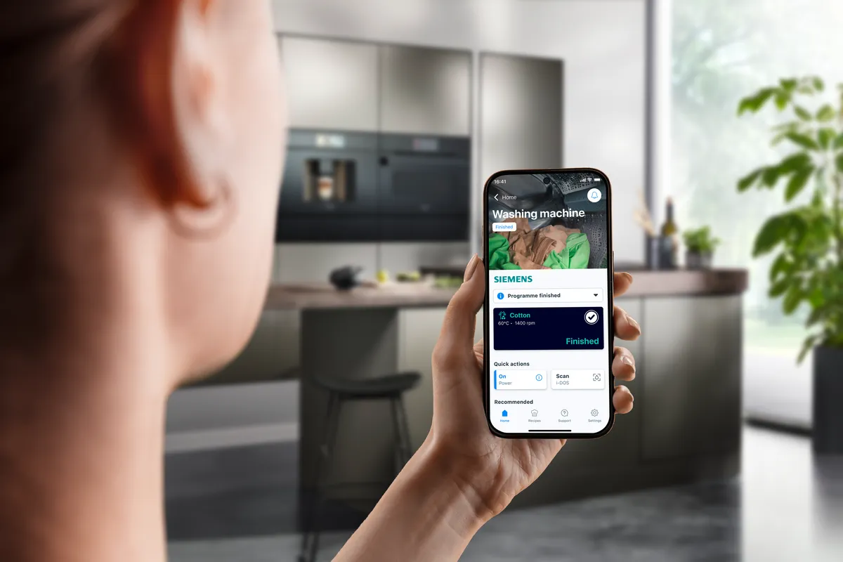 En person holder en smartphone foran et moderne køkken. På skærmen vises en app, der styrer en vaskemaskine og viser, at programmet er færdigt.