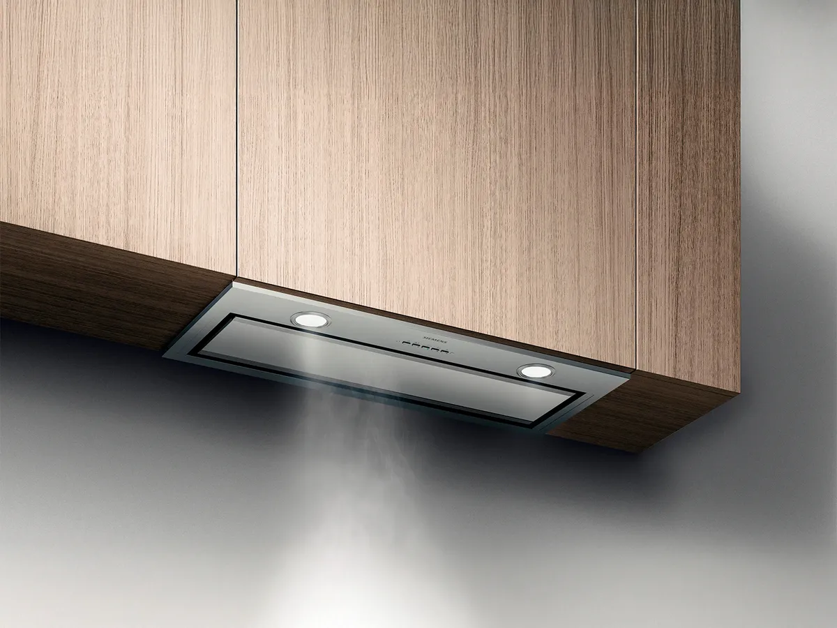 Chaminé de cozinha moderna com design em aço inoxidável e vidro, montada sob armários de madeira, emitindo vapor da cocção abaixo.