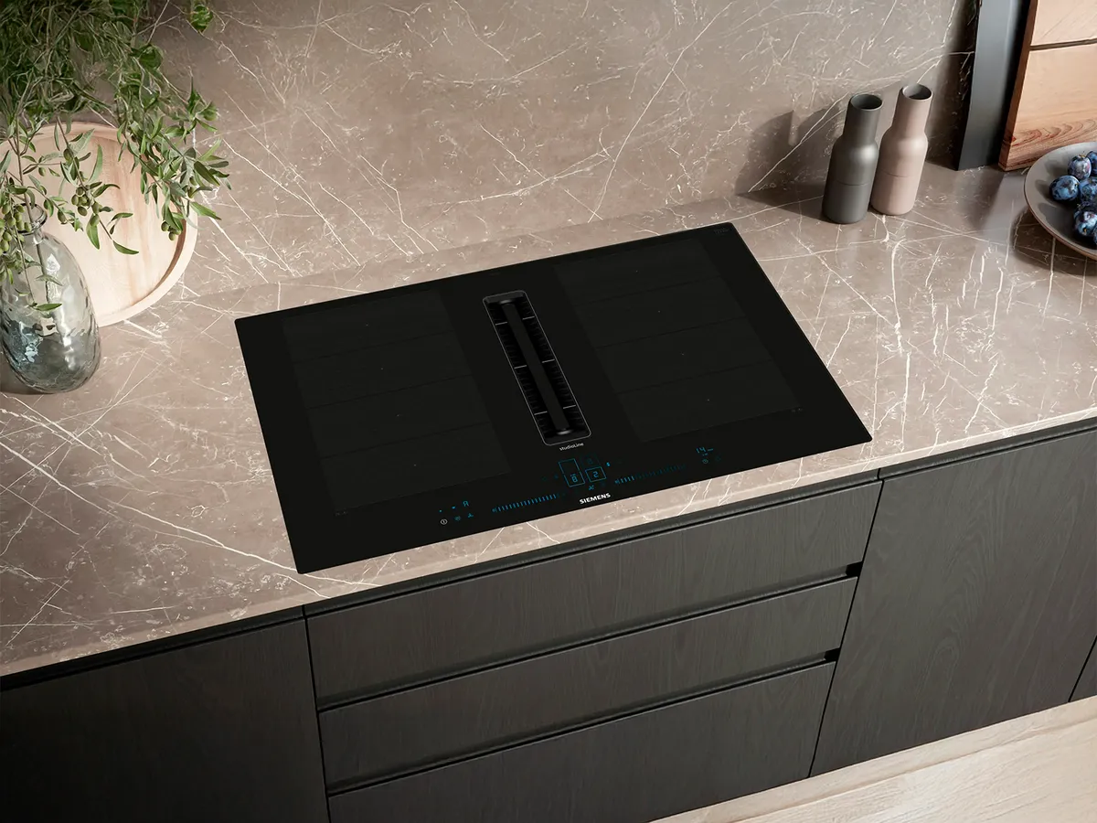 Modern svart induktionshäll monterad i en ljus marmorbänk, med köksfläkten integrerad i mitten av hällen och en stilren design med köksaccessoarer i närheten.