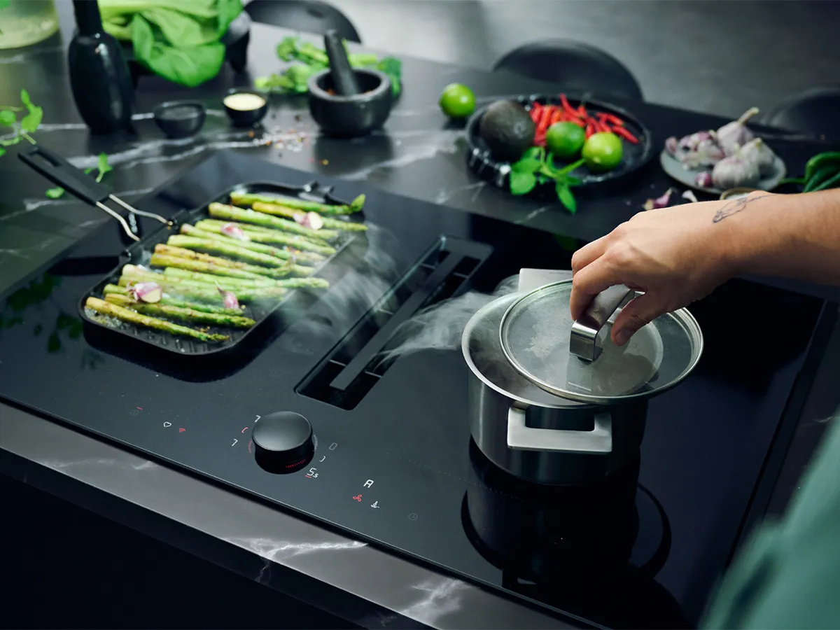 A hand lifts the lid of a steaming pot while grilled asparagus sizzles on a sleek, modern stovetop adorned with fresh ingredients.
