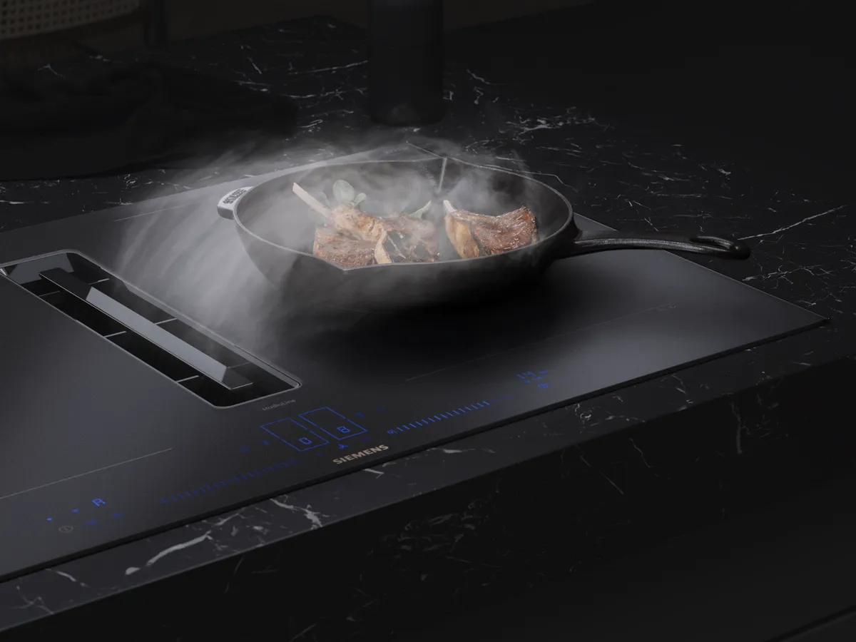 Uma placa de ventilação com um acabamento mate e fumo a sair de uma frigideira que cozinha carne e legumes.
