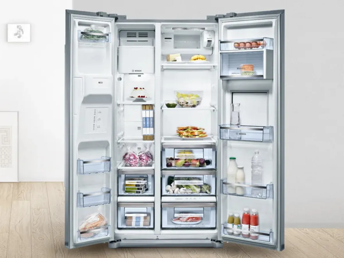 A stainless steel refrigerator stands in a modern kitchen beside a countertop with green limes and a bowl of fruit.